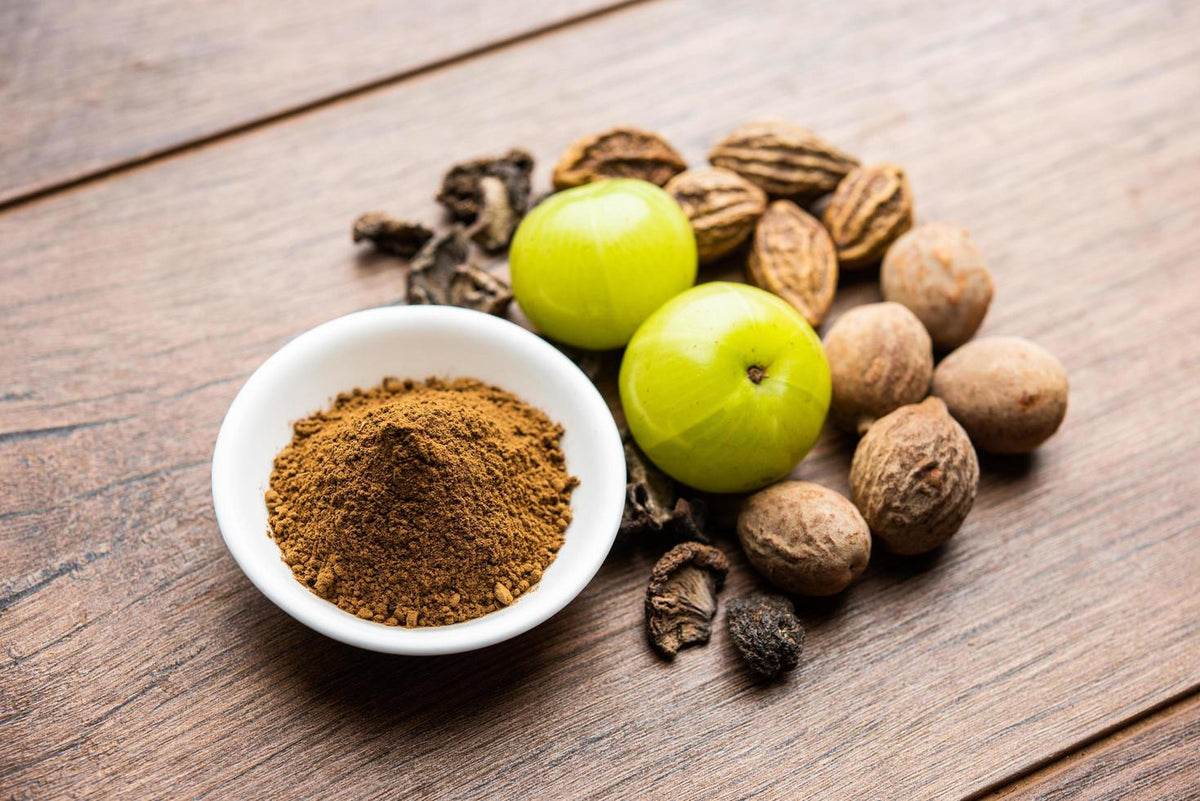 Triphala powder in white bowl with dried fruits and roots beside it